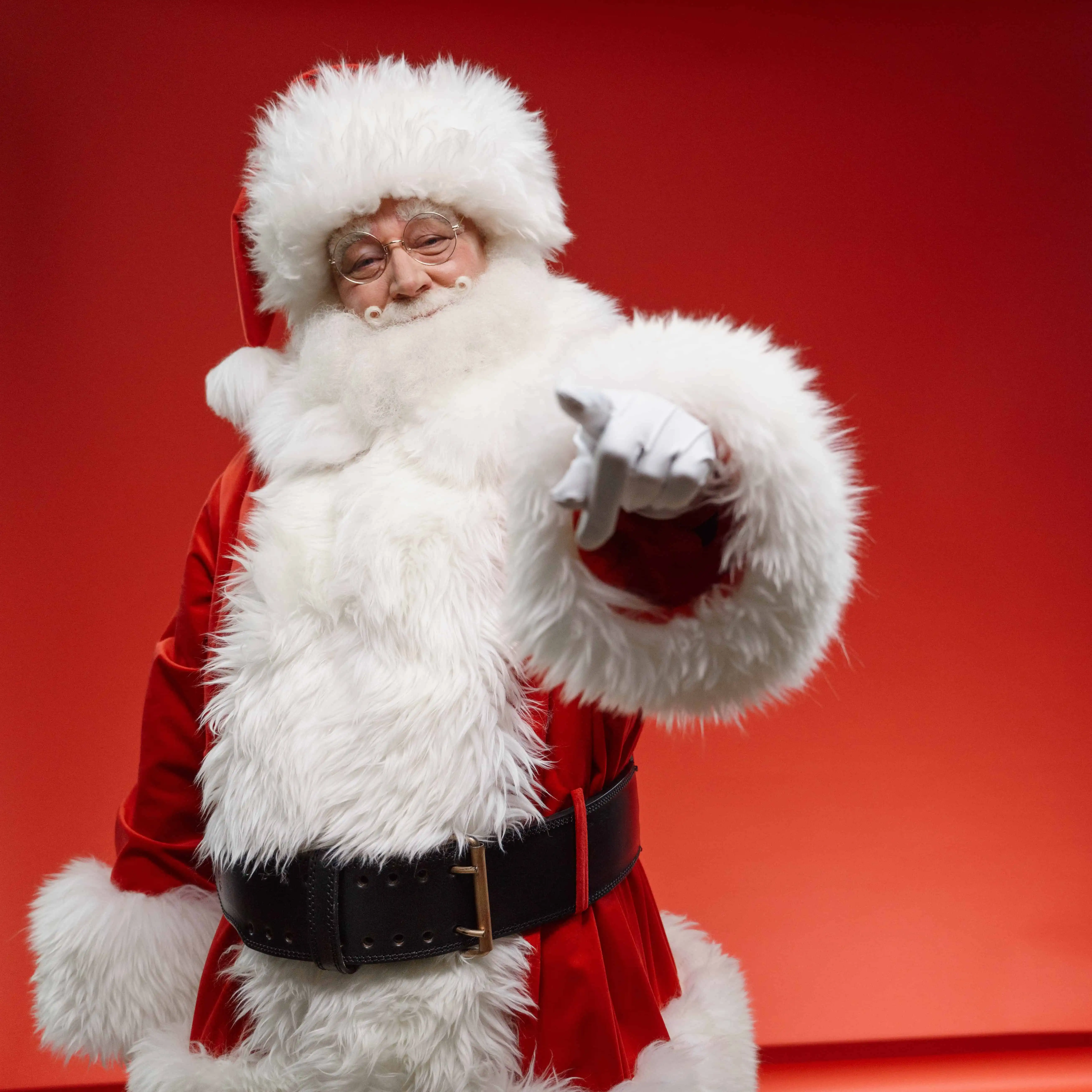 A picture of Santa smiling and standing in front of a red wall, pointing. 