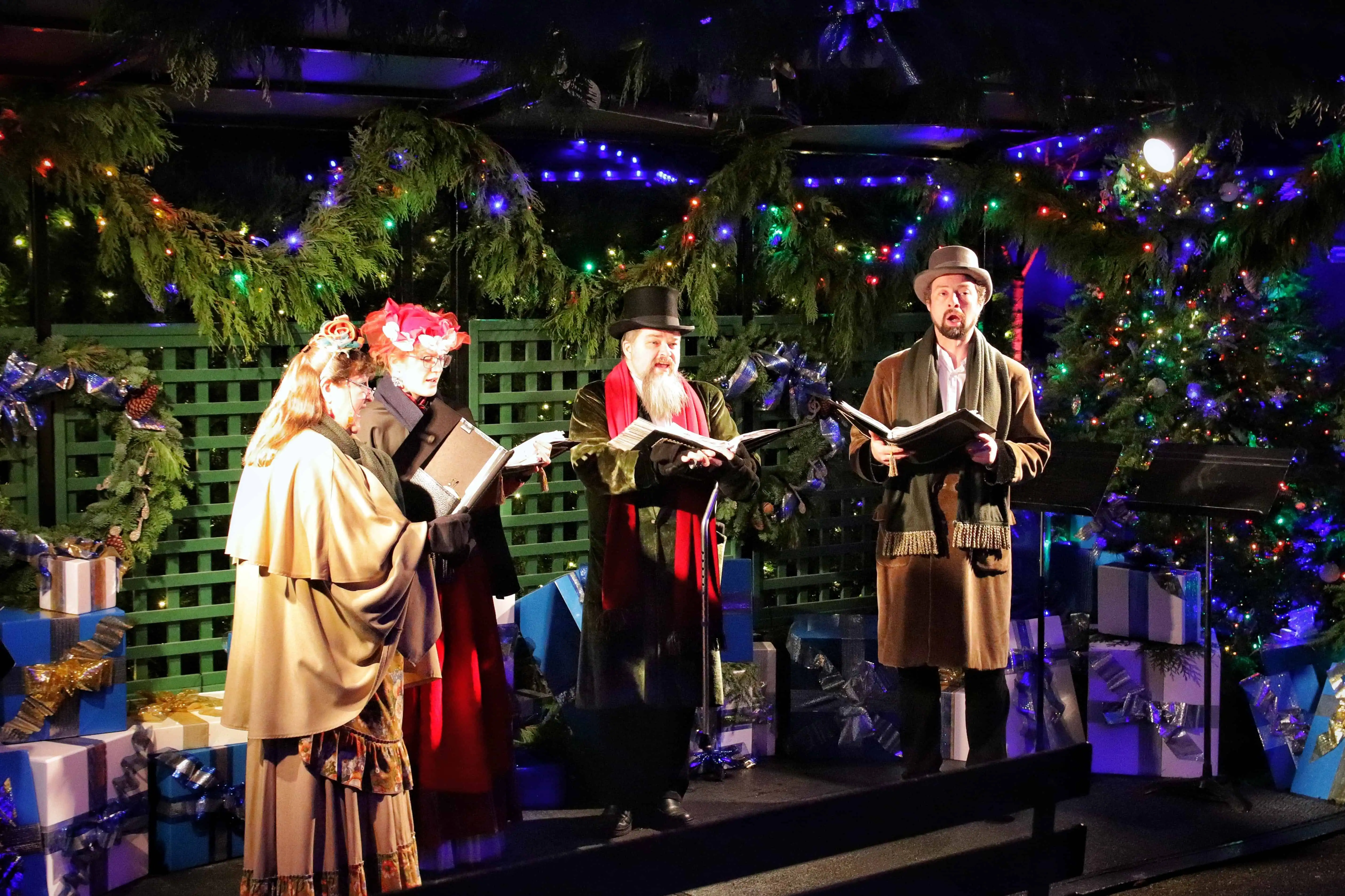 A picture of carolers with presents, lights, and Christams decor behind them.